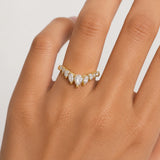 A gold ring with marquise-cut and round clear stones set in a V-shape design, shown worn on a hand against a light gray background.